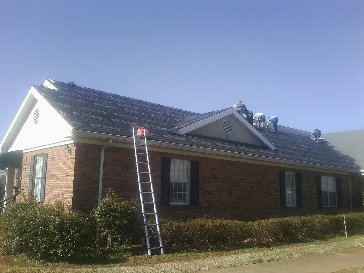Crew tearing shingles off a brick ranch roof with a ladder set — Metal Roof Repair work by our crew in Monson