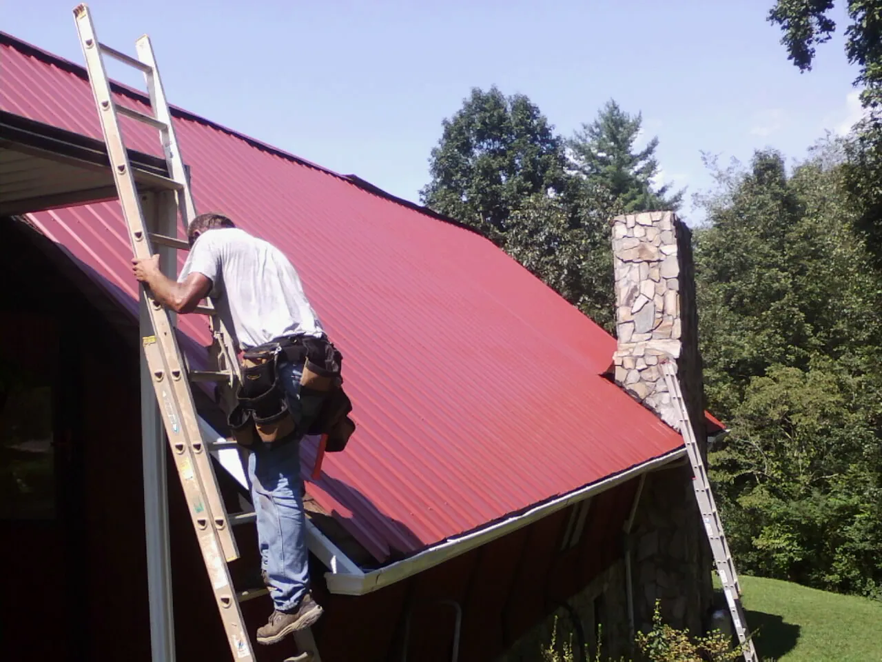 Roofer on a ladder beside a red metal roof and stone chimney — Soffit & Fascia Repair by Roofing Brothers in Monson